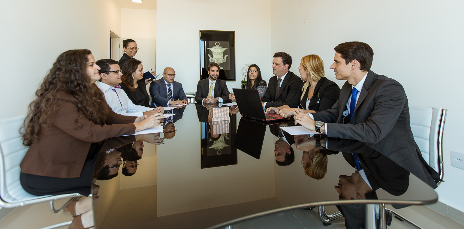 Homens e mulheres, vestidos com roupas formais, reunidos ao redor de uma mesa de reuniões.