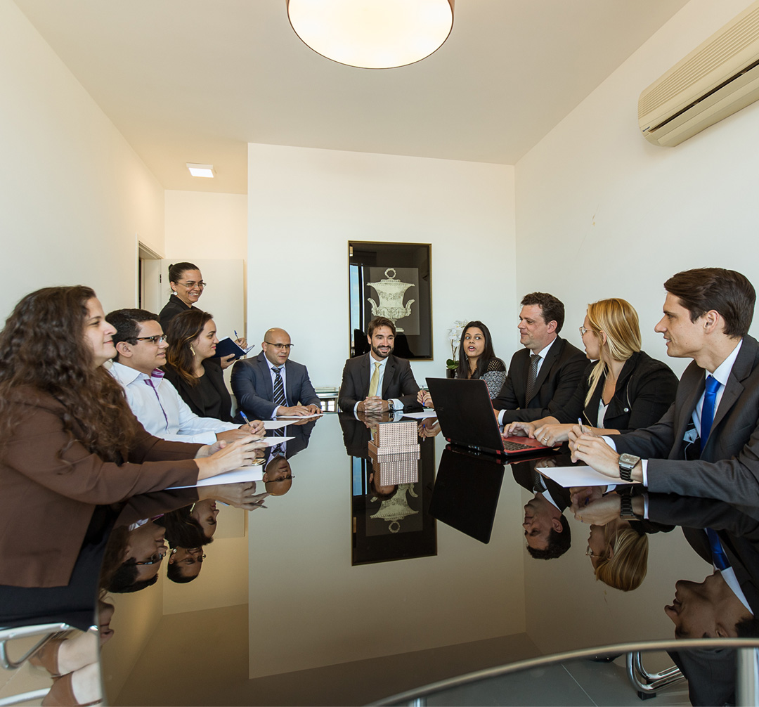 Homens e mulheres, vestidos com roupas formais, reunidos ao redor de uma mesa de reuniões.